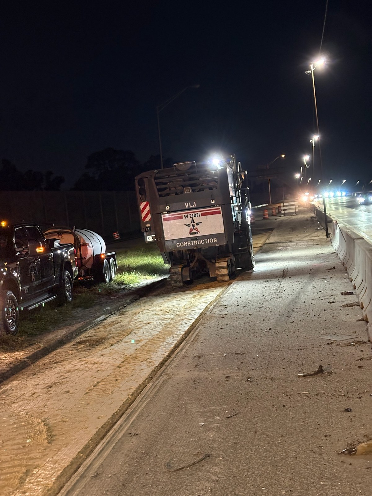A VLJ Construction W 210Fi milling machine working a highway lane at night behind a concrete barrier wall.